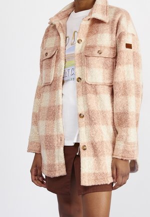 Personne portant une veste à boutons en tissu duveteux à carreaux rose et crème, par-dessus un t-shirt blanc imprimé et une jupe marron avec une fermeture éclair frontale.