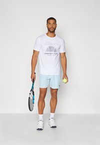 Balta sportinė marškinėlė su "ellesse" logotipu, šviesiai mėlynos šortai, laiko teniso raketę ir kamuoliuką, dėvi baltus sportbačius ir apačios kojines.