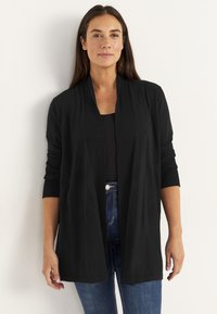 Frau mit langen, dunklen Haaren, die eine schwarze Cardigan mit offenem Vorderteil über einem schwarzen Oberteil und blauen Jeans trägt, vor einem schlichten weißen Hintergrund.