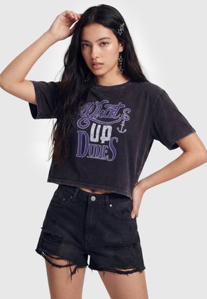 Unge kvinde med langt mørkt hår iført stjerneformede øreringe og hårklemmer, en sort cropped T-shirt med tekst og sorte revnede denimshorts.