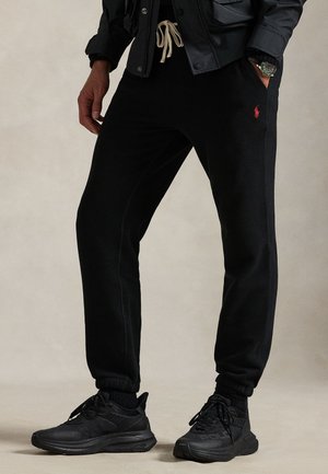 Personne portant un pantalon de jogging noir avec un logo rouge, des baskets noires et une veste sombre, les mains dans les poches.