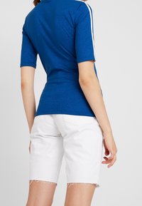 Blå kortärmad topp med en glänsande textur, med vita ränder längs axlarna. Kombinerad med vita fransade shorts.