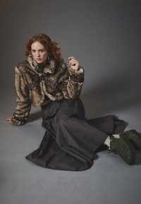 Faux fur cropped jakke med høy krage og brune lærremmer, kombinert med et langt svart skjørt og mørkegrønne ankelstøvler.