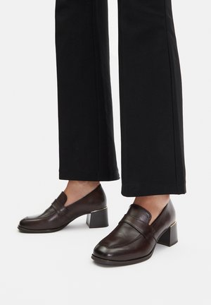 Braune Leder-Blockabsatzschuhe mit abgerundeter Spitze, Schlupfdesign und goldenem Akzent an der Ferse, kombiniert mit schwarzer Hose.