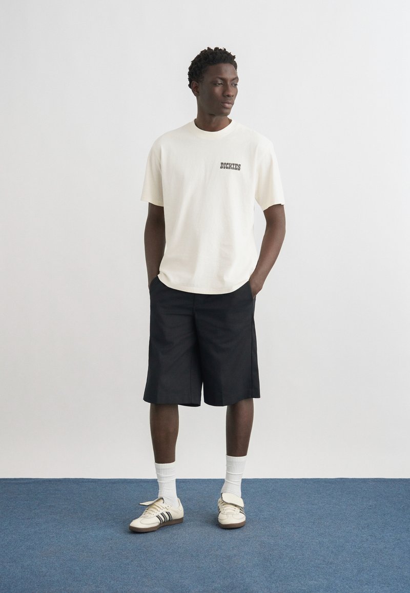 Uomo in piedi con le mani in tasca, indossa una maglietta bianca Dickies, pantaloncini neri fino al ginocchio, calzini bianchi e sneakers bianche su un pavimento blu.