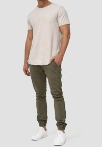 Beige T-Shirt mit kurzen Ärmeln und Brusttasche, kombiniert mit olivgrünen, zulaufenden Jogginghosen mit elastischen Bündchen und weißen Sneakern.