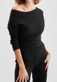 Pull noir côtelé à épaules dénudées avec manches longues et une large ceinture, présentant un design texturé et une silhouette ajustée.