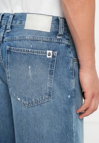 Vaqueros de mezclilla azul con un lavador desgastado; presenta un parche de cuero blanco en la parte posterior y manchas de pintura en la tela. Incluye una etiqueta con el logo.