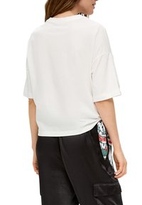 Vrouw in een losse witte T-shirt met korte mouwen en zwarte broeken, met een kleurrijke gestructureerde sjaal gestrikt op de heup, gezien vanuit de rug.