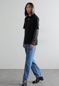 Svart T-shirt med kort ärm och ett cirkulärt logotyp, över en långärmad mönstrad skjorta, tillsammans med ljusblå raka jeans och svarta skor.