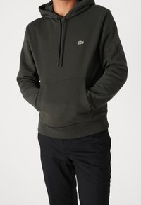 Felpa con cappuccio verde scuro con tasca a marsupio e coulisse sul cappuccio. Presenta un piccolo logo ricamato sul petto a sinistra. Abbinata a pantaloni neri.