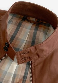 Chaqueta marrón con cierre de cremallera, botones negros y forro de tartán en tonos beige, marrón y gris. Textura exterior lisa, cuello estructurado.