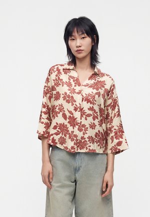 Femme debout devant un fond uni, portant une chemise beige à motifs floraux rouges et un jean bleu clair, regardant l'appareil photo.