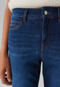 Dunkelblaue Jeans aus Denim mit glatter Textur, die dezente Auswaschungen, Kontrastnähte und einen Knopfverschluss an der Taille aufweisen.