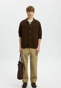 Brązowa koszula z guzikami i dwoma kieszeniami na piersi, zestawiona z beżowymi spodniami w szerokim kroju i brązowymi butami, trzymając pasującą brązową torbę na ramię.