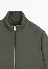 Giacca verde scuro con collo alto e cerniera frontale argento, esposta su uno sfondo bianco semplice.