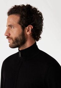 Crna zip-up dukserica s visokim ovratnikom, izrađena od glatkog materijala, s uskim krojem i suptilnim detaljem šivanja na zipu.
