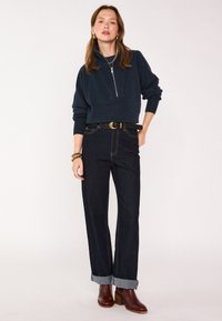 Maglione blu navy con zip e polsini a coste, abbinato a jeans scuri a vita alta con orli arrotolati e una cintura nera con fibbia dorata.