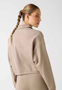 Jasno beżowa skrócona bluza dresowa o luźnym kroju, miękkiej fakturze i kołnierzowym dekolcie. Posiada opuszczone ramiona i minimalne detale przeszyć.
