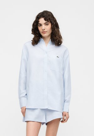 Donna con capelli ricci che indossa una camicia azzurro chiaro abbottonata e pantaloncini coordinati, in piedi davanti a uno sfondo bianco uniforme.