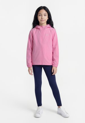 Jong meisje staat en draagt een roze capuchonjas, marineblauwe legging en witte sneakers op een effen witte achtergrond.