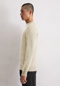 Cremfarbener Pullover mit geripptem Saum und Bündchen, strukturiertem Stoff, Rundhalsausschnitt, taillierter Silhouette, getragen über schwarzen Hosen.