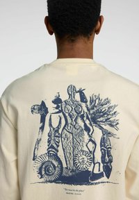 Krämfärgad långärmad skjorta med en blå grafik av abstrakta figurer och artefakter på baksidan, med texten "Det här måste vara platsen."