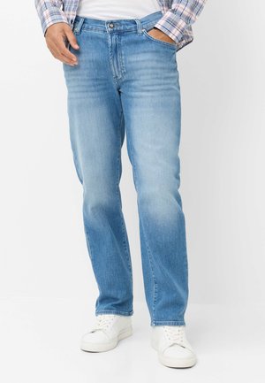 Mand iført lyseblå jeans, hvide sneakers, en hvid skjorte og en ternet skjorte med én hånd i lommen mod hvid baggrund.