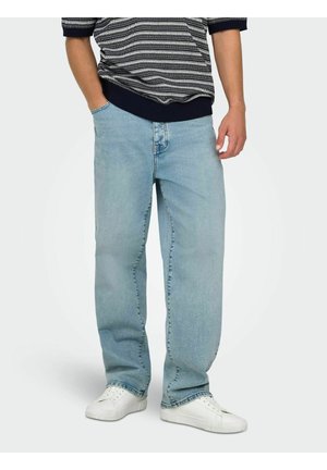 Osoba nosząca jasnoniebieskie, luźne dżinsy, białe trampki i krótki rękaw w czarno-biały wzór, stojąca z jedną ręką w kieszeni.