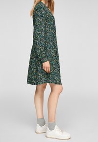 Blommig klänning i mörk teal med långa ärmar och avslappnad passform, med flerfärgade blommönster. Matchad med vita sneakers.