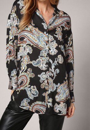 Femme portant une chemise noire ample boutonnée avec des motifs cachemire multicolores et un pantalon en cuir noir, montrée du cou au milieu de la cuisse.