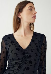Čierny vzorovaný top so V-čkovým výstrihom a dlhými priesvitnými rukávmi. Tkanina má kvetinovú textúru s nariasením v oblasti poprsia.