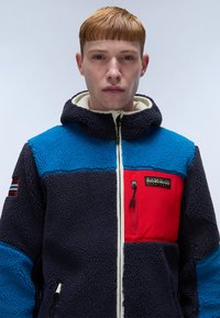 Fleece jacket featuring a blue, navy, and red color block design, with a zippered chest pocket and brand logo on the front. Textured fabric.