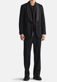 Completo nero su misura con colletto sciallato fluente, sciarpa coordinata e pantaloni slim-fit. Tessuto testurizzato con leggero lucido; finitura liscia.