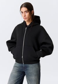 Schwarzer Kapuzenpullover mit Reißverschluss, großer Kapuze, lockerem Schnitt, Fronttaschen und gerippten Bündchen, kombiniert mit hellblauen Jeans.