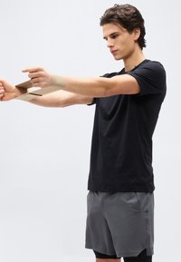 Νέος άνδρας με μαύρο πουκάμισο και γκρι σορτς τεντώνει μια μπεζ λωρίδα αντίστασης με τα χέρια τεντωμένα μπροστά σε απλό φόντο.