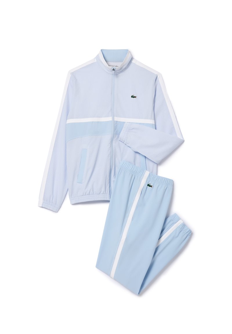 Ensemble de sport bleu clair fabriqué en tissu respirant. Comprend des bandes blanches, une veste zippée et un pantalon avec ceinture élastique. Détail du logo Lacoste.