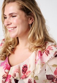 Bloes met bloemenprint in lichtgewicht stof, versierd met roze en bordeaux bloemen, met een gerimpelde halslijn en subtiele glans. Krullend blond haar met zichtbare oorbellen.