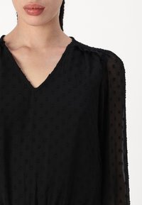 Blouse noire avec un col en V, en tissu texturé avec des points en relief, manches longues en voile, et détails froncés aux épaules.
