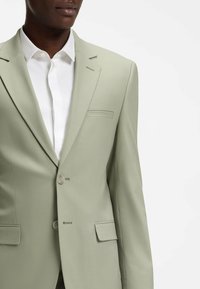 Muž v světle zeleném saku přes bílou košili s límečkem, zobrazený od ramen po střed trupu na jednobarevném světlém pozadí.