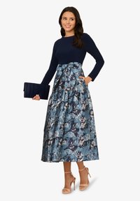 Maxi-Kleid mit einem navyblauen, langärmeligen Oberteil und einem strukturierten, blau mit Blumenmuster bedruckten Rock. Ein Schleifendetail an der Taille und eine schlanke, dunkle Clutch runden das Outfit ab.