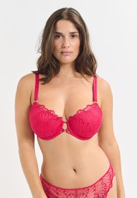 Sujeto rojo de encaje con bordes festoneados, soporte de aros y herrajes decorativos en el centro. Tirantes delgados ajustables y detalle de patrón floral.