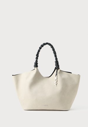 Beige DKNY tote bag met zwarte gevlochten handvatten en een zilveren logo charm, weergegeven tegen een witte achtergrond.