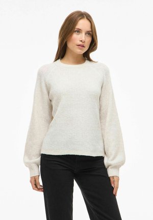 Pull beige clair avec un motif texturé, encolure ronde et manches bouffantes. Porté avec un pantalon noir, avec des poignets et un ourlet côtelés.