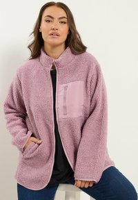 Chaqueta de forro polar en rosa suave con un bolsillo en el pecho con cremallera, cuello alzado y bolsillos laterales, con una superficie texturizada y esponjosa.