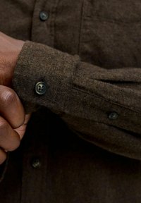 Dark brown wool shirt cuff with black buttons, worn by a person adjusting the sleeve with their hand.