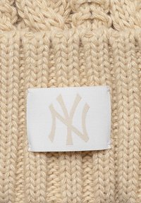 Bežinė mezginio medžiaga su tekstūruotu raštu, turinti baltą etiketę su bežiniu "NY" logotipu, siuvinėtu ant jos.