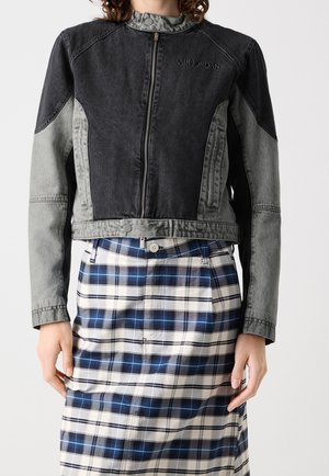 Osoba ubrana w czarno-szarą kurtkę Air Jordan na zamek błyskawiczny oraz niebieską, białą i czarną kratowaną spódnicę, stojąca na jednolitym tle.