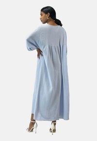 Abito lungo di cotone blu chiaro con vestibilità ampia, maniche larghe e dettagli arricciati sulla schiena; abbinato a sandali con tacco alto bianchi.