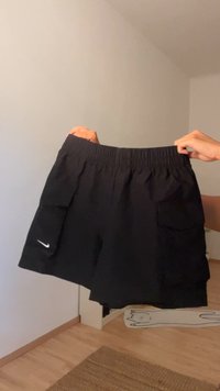 Rankos laiko juodus Nike šortus su elastinga juosta ir kišenėmis kambaryje su šviesiais mediniais grindimis ir baltais sienomis.